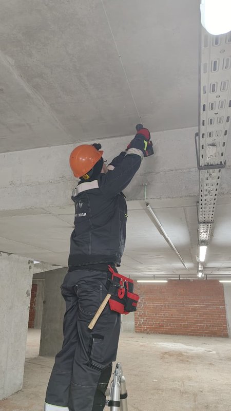 Монтажник в каске крепит кабельный лоток к бетонному потолку на объекте